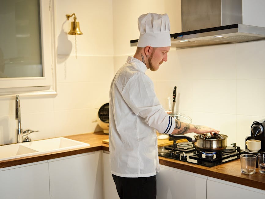 A tattooed chef wearing a hat cooks in a modern kitchen, focusing on a pot on the stove
