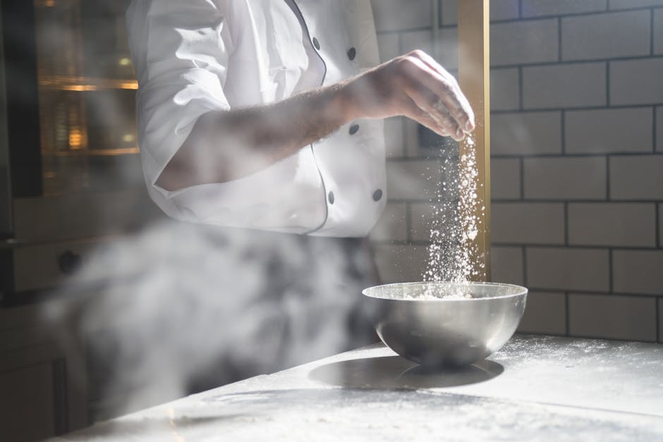 Chef in white uniform sprinkling flour into a steel bowl in a modern kitchen