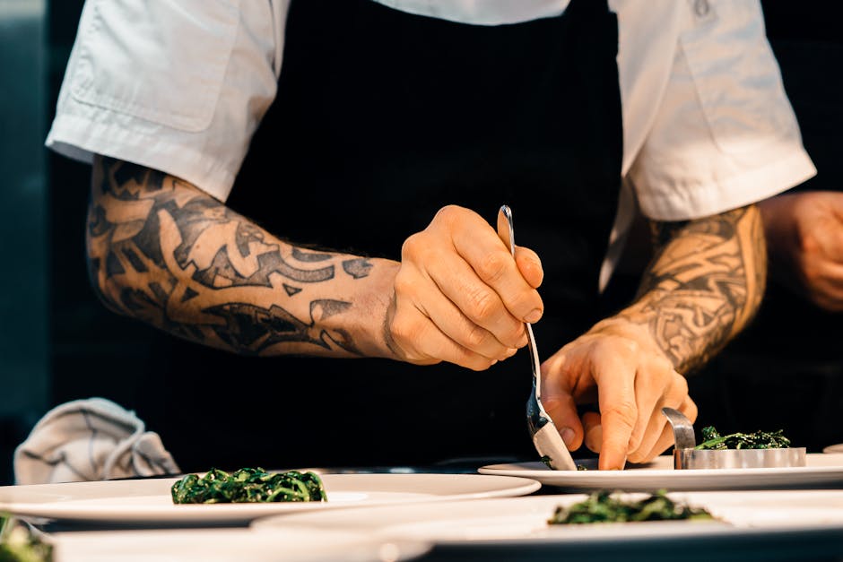 A tattooed chef carefully plates a spinach dish in a professional kitchen setting, showcasing culinary expertise